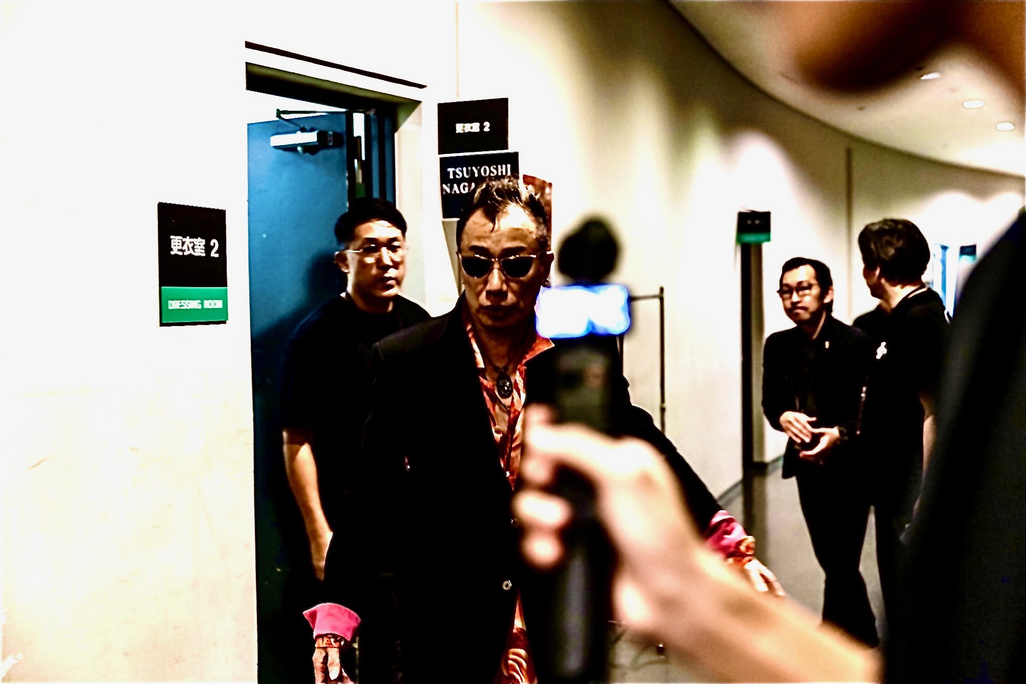 廊下でインタビューされるサングラス男性